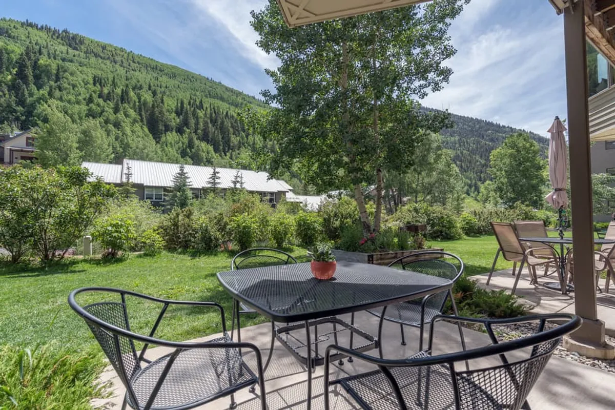 A tranquil outdoor patio area with a metal dining table set amidst lush greenery, framed by impressive mountain views in the background. The patio is surrounded by plants and trees that evoke a feeling of privacy and direct connection to nature.