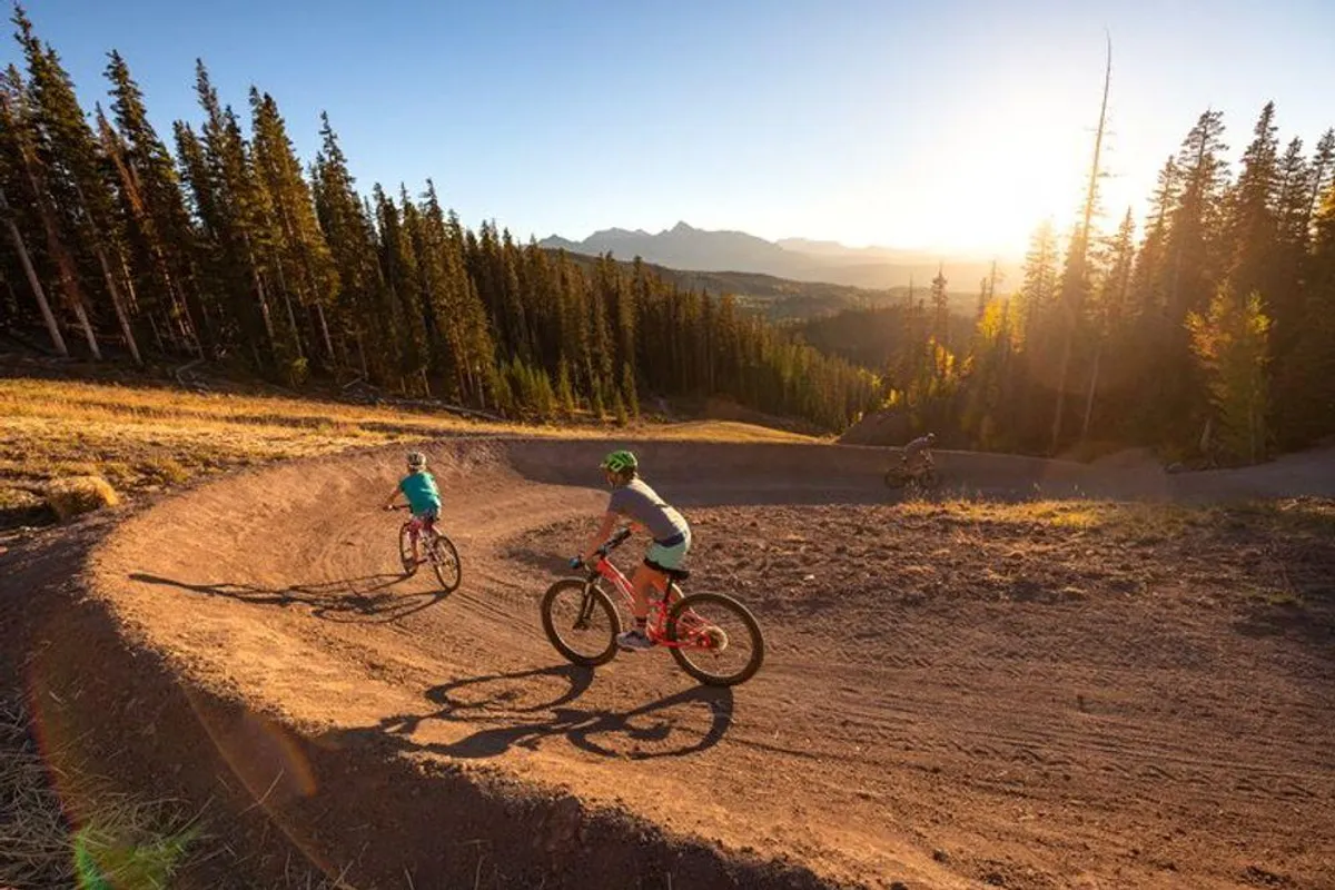 Mountain bikers navigate a sunset-lit curved trail through pine trees, demonstrating the area's outdoor adventure opportunities. The image captures the excitement and accessibility of biking and hiking from the property.