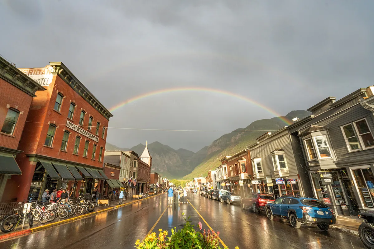 A vibrant street scene in Telluride featuring historic buildings, a rainbow arching over wet streets, and people walking or biking. Local shops and cafes are visible, capturing the charm and walkability of the neighborhood.