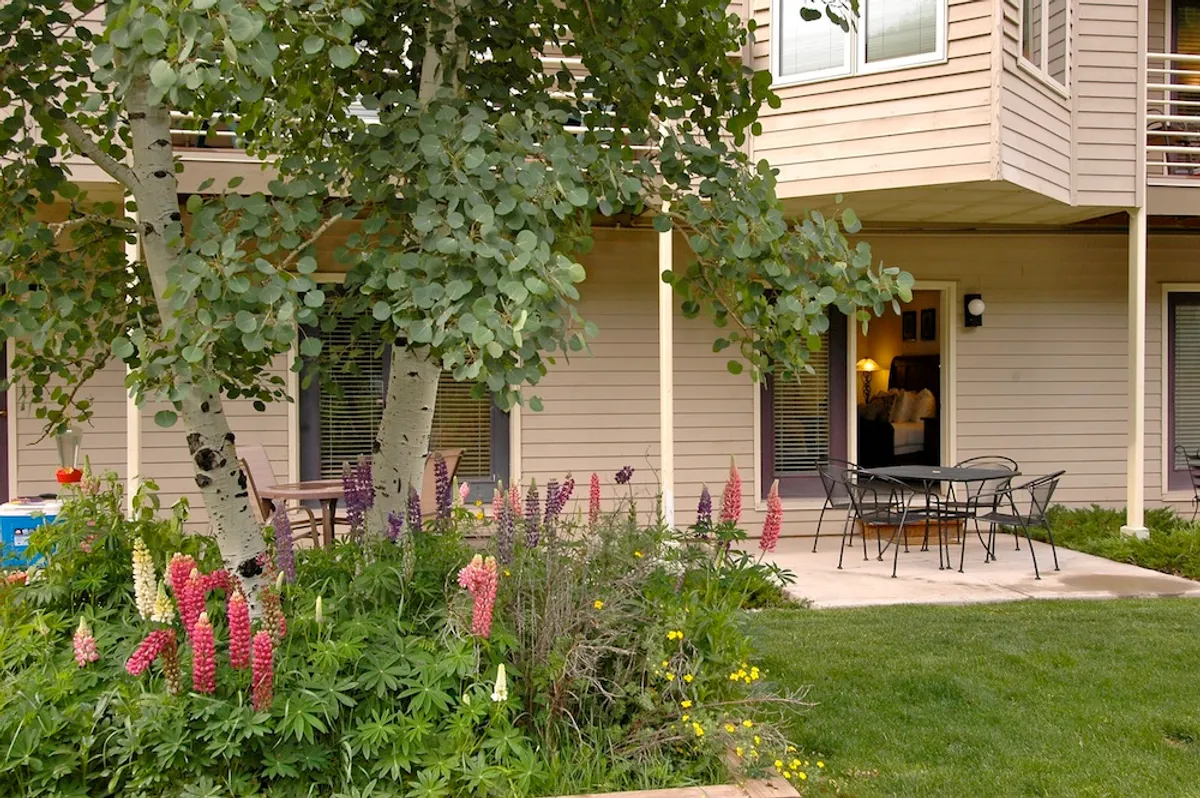A tranquil outdoor seating area set among colorful flowers and a shady tree. The space is designed for relaxation, with tables and chairs inviting groups to gather for meals or conversation. The riverside vibe is enhanced by the lush landscaping and open sky.