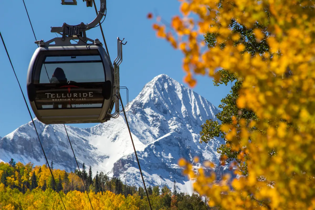 A gondola glides above vibrant autumn foliage with a dramatic mountain peak in the background. The scene naturally evokes adventure and romance with a sense of local color and seasonal beauty.