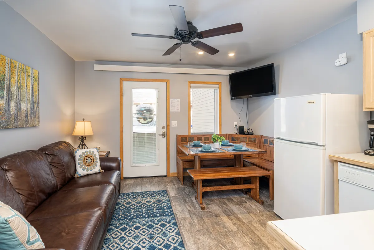A warm, inviting interior featuring a brown leather couch, a wooden dining table, decorative wall art, and kitchen appliances in the background. The layout demonstrates seamless transitions between lounging, dining, and cooking spaces.