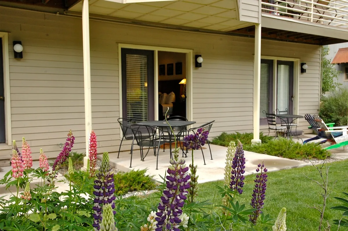 A flower-filled garden with comfortable patio furniture arranged invitingly near entrance doors. The area is bathed in natural light, suggesting the perfect spot for both relaxing and social gatherings outside.