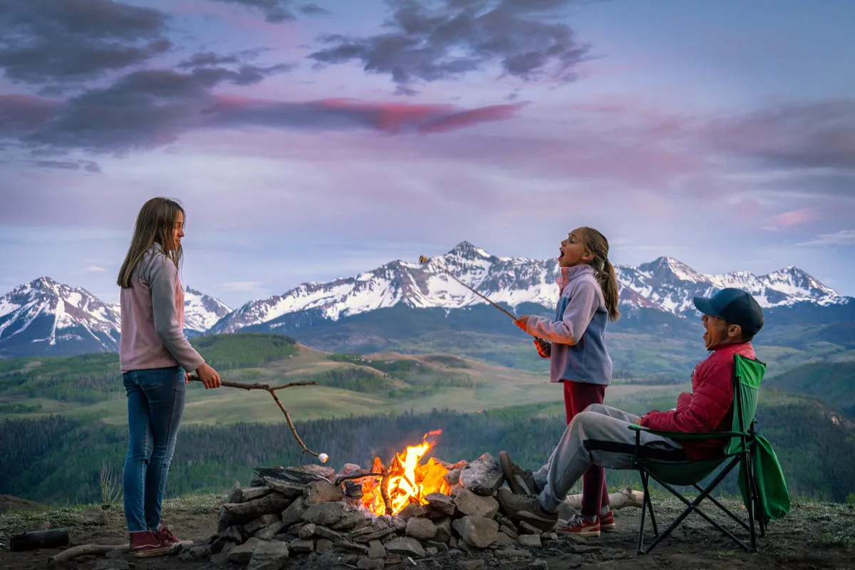 A group of children roast marshmallows around a campfire with mountains in the background. A man sits in a chair nearby, watching the children. The setting sun creates a warm, inviting glow, and the mood is lively and family-oriented, emphasizing shared experiences and outdoor fun.