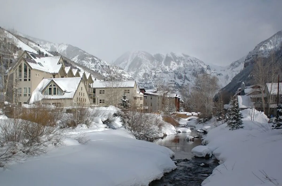 Snow-covered mountains loom in the background while a gentle stream runs through the property, with distinct architectural details of the lodge visible. This setting evokes both winter adventure and a tranquil riverside escape.
