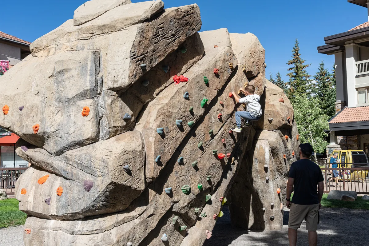A child climbs a rock wall outdoors while an adult observes, surrounded by greenery and open space. The scene is energetic, playful, and highlights hands-on family bonding experiences. The activity builds confidence and encourages outdoor play.