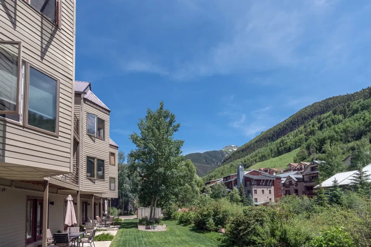A tranquil exterior view of a green lawn in front of the building, outdoor seating areas, and a backdrop of majestic mountains. The scene is calm and inviting, perfect for relaxing, social gatherings, and watching sunsets. The environment is safe and friendly, suitable for family activities.