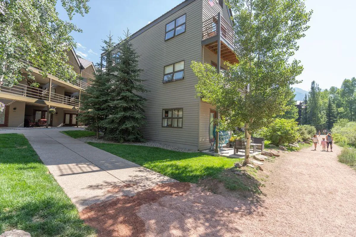 This exterior shot highlights the building nestled among lush greenery, with a walkway and people strolling. The image conveys accessibility to trails and town, suggesting the ease of exploring the area on foot.