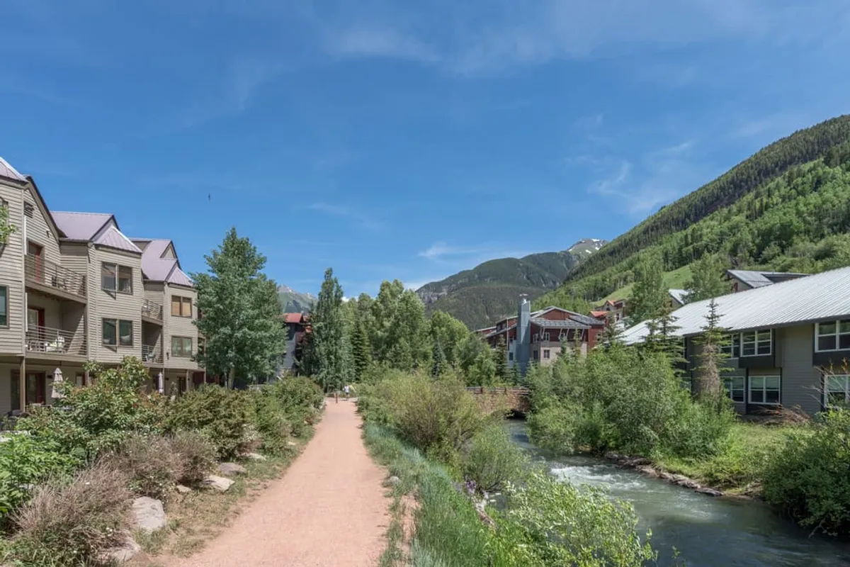 A scenic walkway runs alongside a gentle stream with green foliage on both banks, leading toward nearby buildings. The path is inviting, with sunlight dappling the trail and mountains rising in the distance, suggesting immediate access to nature.