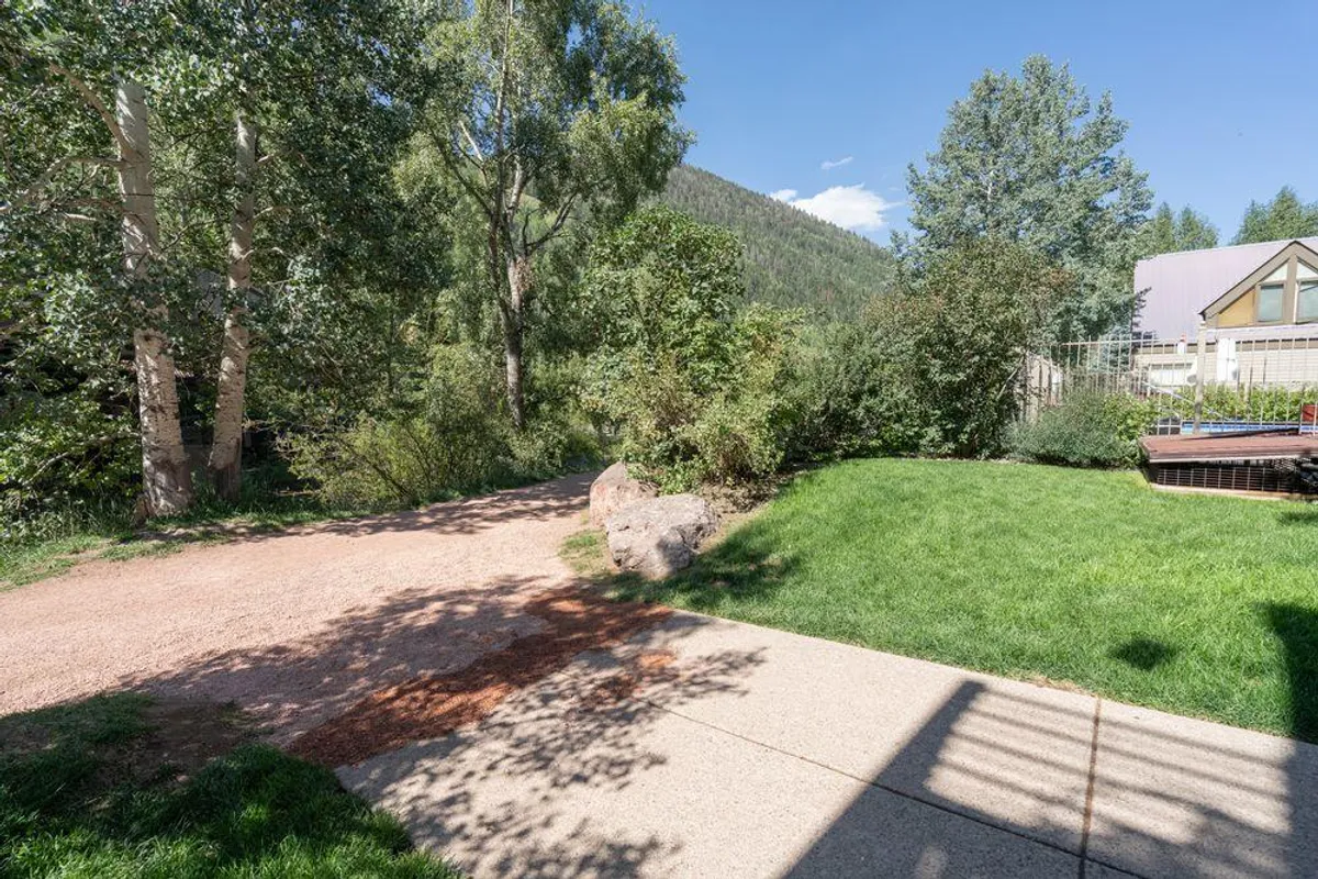 A tranquil grassy area sits next to a winding pathway, flanked by natural rock formations and framed by distant trees and mountain views. The space invites exploration, with guests visible walking or relaxing in the sunshine, taking in the serene environment.