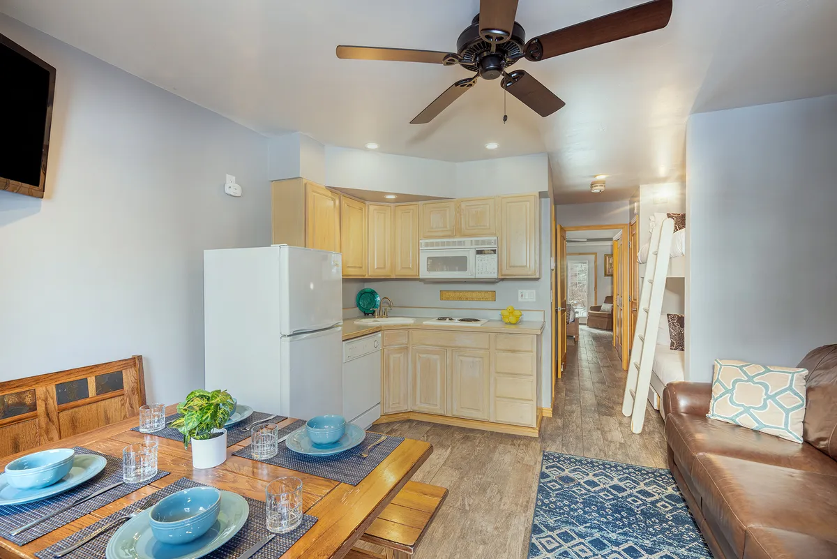 This image captures an open-plan space featuring a dining table set with dishes, visible kitchen appliances, and a living area with a comfy couch. Bunk beds are present in the frame, but the primary focus is on the communal cooking, dining, and relaxing zones.